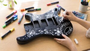 黒いギターボディに白いペイントマーカーで花や星の一筆描きアートを描いている手元