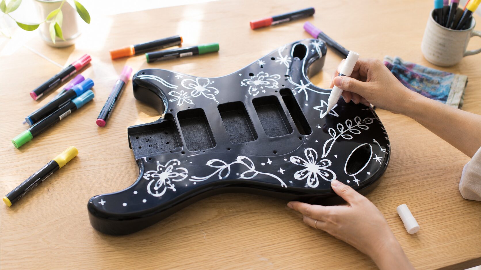 黒いギターボディに白いペイントマーカーで花や星の一筆描きアートを描いている手元